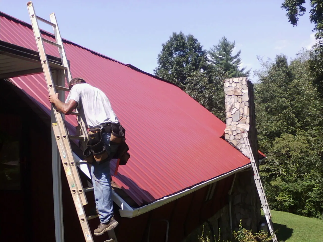 Roofer on a ladder beside a red metal roof and stone chimney — Soffit & Fascia Repair by Roofing Brothers in Woodsville